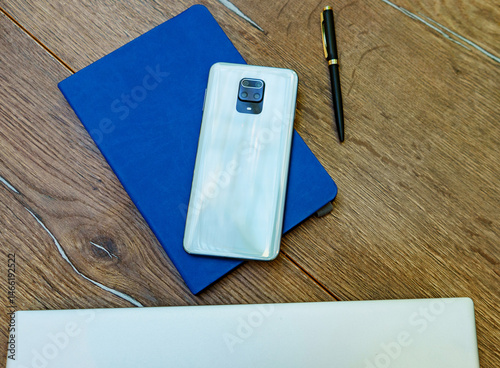 Top view of a wooden desk with a smartphone, a closed blue notebook, a pen, and a silver laptop. The modern phone and office supplies suggest a digital professional workspace. 