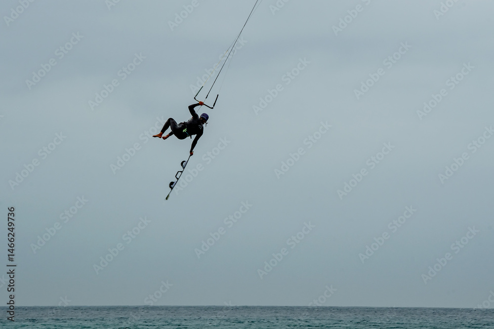 Fototapeta premium Kite surfers in the waves on a windy day. Tropical beach huts and spectators watching on the sandy beach, Bright colourful kites fill the cloudy sky. Turquoise water. copy space. Sport vacation.