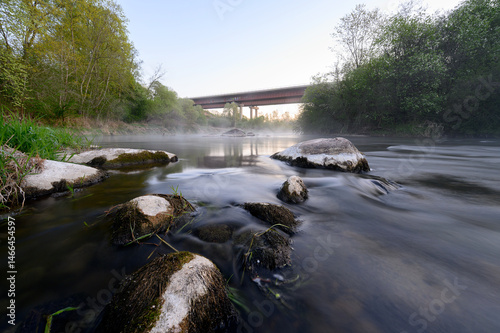 Obraz na płótnie Sunrise over a misty river surrounded by lush green trees in spring. Tranquil nature landscape with calm water, fog, and golden sunlight reflecting on the surface. Ideal for eco, travel, wellness. w salonie