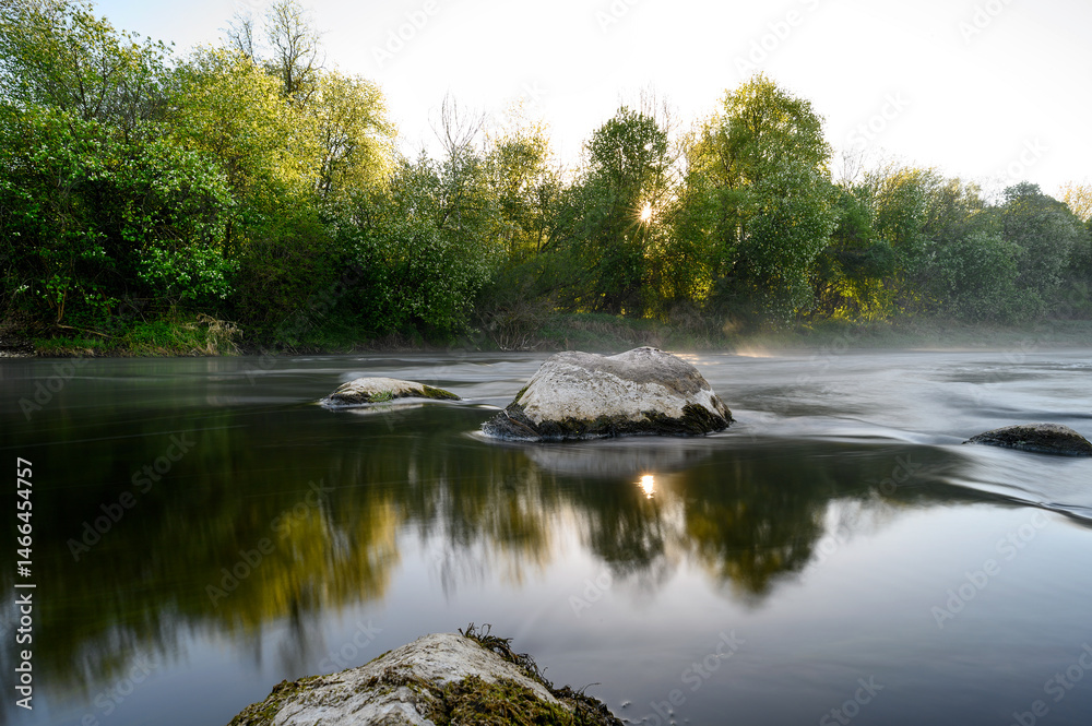 Sunrise over a misty river surrounded by lush green trees in spring. Tranquil nature landscape with calm water, fog, and golden sunlight reflecting on the surface. Ideal for eco, travel, wellness.