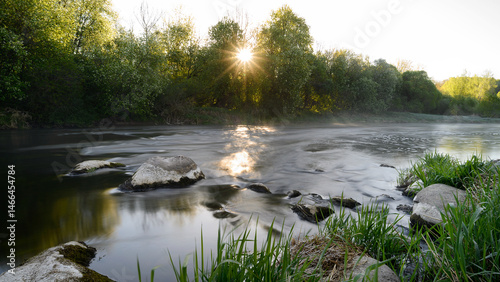 Obraz na płótnie Sunrise over a misty river surrounded by lush green trees in spring. Tranquil nature landscape with calm water, fog, and golden sunlight reflecting on the surface. Ideal for eco, travel, wellness. w salonie
