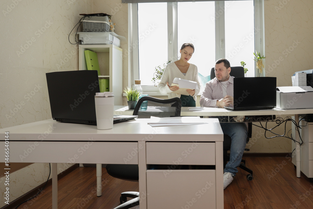 Two colleagues engage in a productive conversation at their shared office desk, with charts and paperwork laid out in front of them, surrounded by office technology and decor