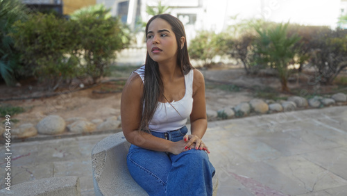 Woman with brunette hair sitting outdoors in an urban park, wearing casual attire, showcasing a serene expression amid daylight and natural surroundings. Free Download Woman with brunette hair sitting outdoors in an urban park, wearing casual attire, showcasing a serene expression amid daylight and natural surroundings. - GFXLoad