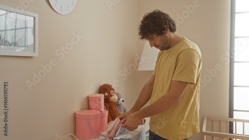 Young hispanic man in bedroom organizing baby clothes with stuffed animals in background while sunlight enters the room, creating a warm interior setting at home