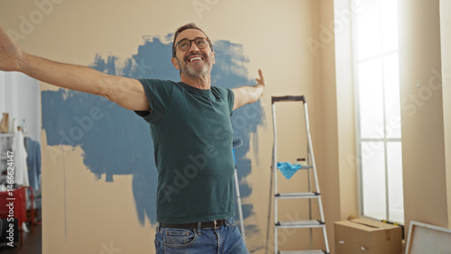 Mature hispanic man smiling with arms outstretched in a newly painted living room, with a ladder and paint supplies, enjoying the fresh feeling of his new home.