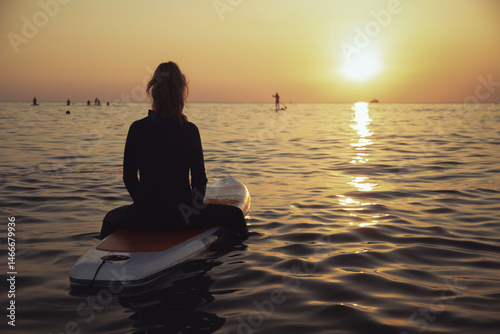 An elderly woman rides paddleboarding in the ocean. Senior female is waiting for a wave on a surfboard. Active old age. Old girl is sitting on a board in the sea at sunset, dressed in a swimming suit
