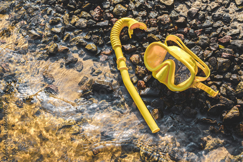 The mask and snorkel lie on a rocky sandy shore near clear water.