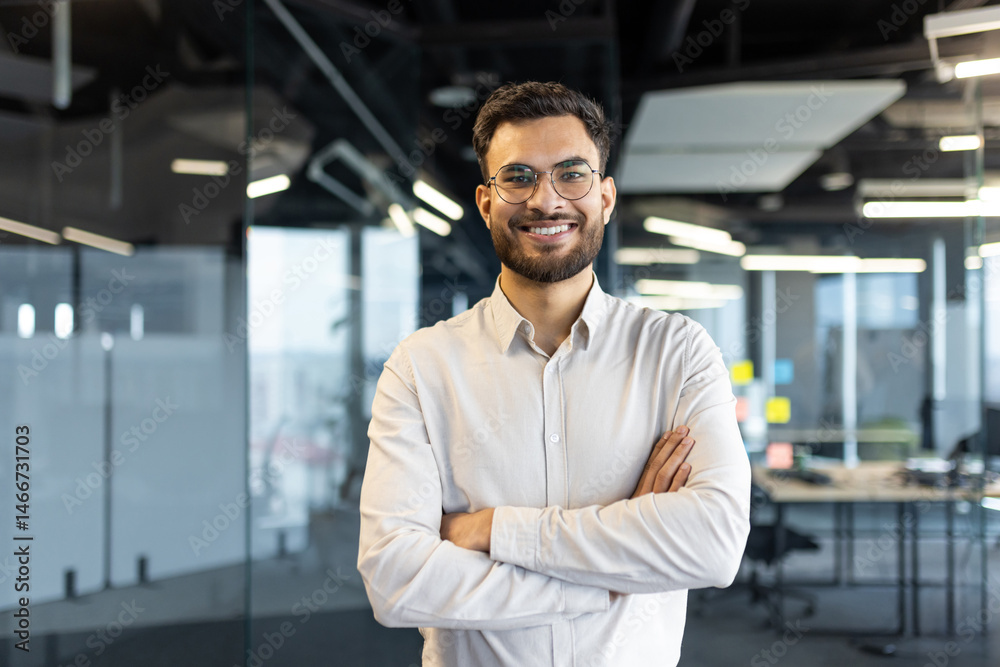 Fototapeta premium Portrait of successful young businessman man smiling and looking at camera with crossed arms. Office worker satisfied with results of achievement at workplace.