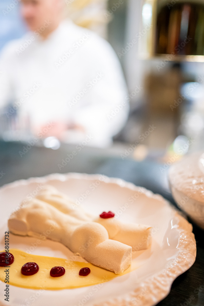 Elegant gourmet dessert on a white plate, shaped like a reclining figure, drizzled with berry sauce and cream. Artful food presentation in a restaurant setting