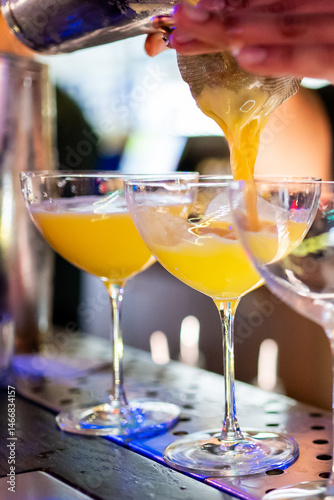 A bartender expertly pours a vibrant yellow cocktail through a strainer into elegant glasses, showcasing the craft of mixology in a dynamic bar setting