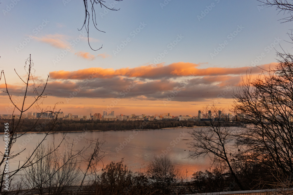 Golden Sunset Reflecting on Calm Waters over Distant City Buildings Framed by Bare Branches in Winter Nature Scene Golden Sunset Reflecting on Calm Waters over Distant City Buildings Framed by Bare Branches in Winter Nature Scene