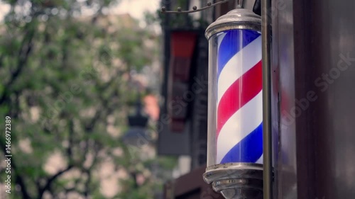 A classic barber pole spins gently in the breeze outside a bustling barbershop, surrounded by urban architecture and greenery.