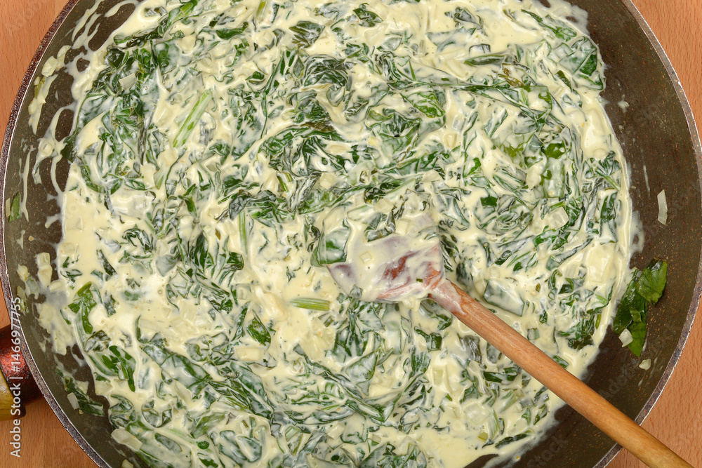 A top-down view shows creamy spinach being stirred in a dark pan with a wooden spoon