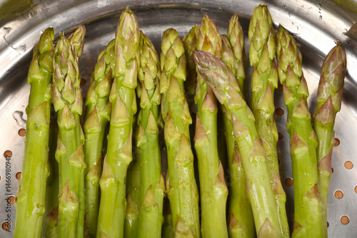 A close-up shows fresh, green asparagus spears arranged inside a metal steamer basket with visible condensation, ready for cooking