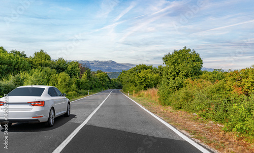 A white sedan drives down a peaceful countryside road flanked by lush greenery and distant mountains under a clear blue sky.
