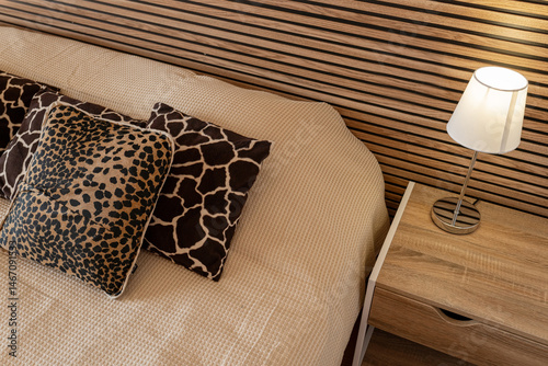 Modern cozy bedroom with a wooden nightstand, lamp, and animal print decorative pillows. Warm and stylish interior with natural wood and neutral tones.