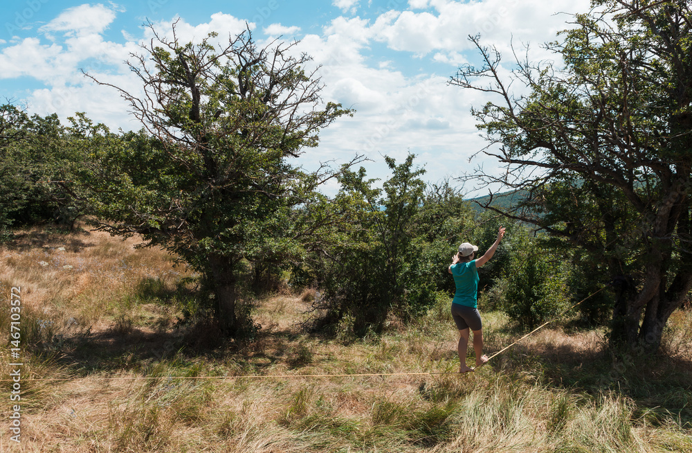 A female slackliner performs acrobatic poses and splits on a slackline stretched between trees on a mountain hilltop in Georgia. Aerial views, dry summer grass, and scenic peaks surround her A female slackliner performs acrobatic poses and splits on a slackline stretched between trees on a mountain hilltop in Georgia. Aerial views, dry summer grass, and scenic peaks surround her