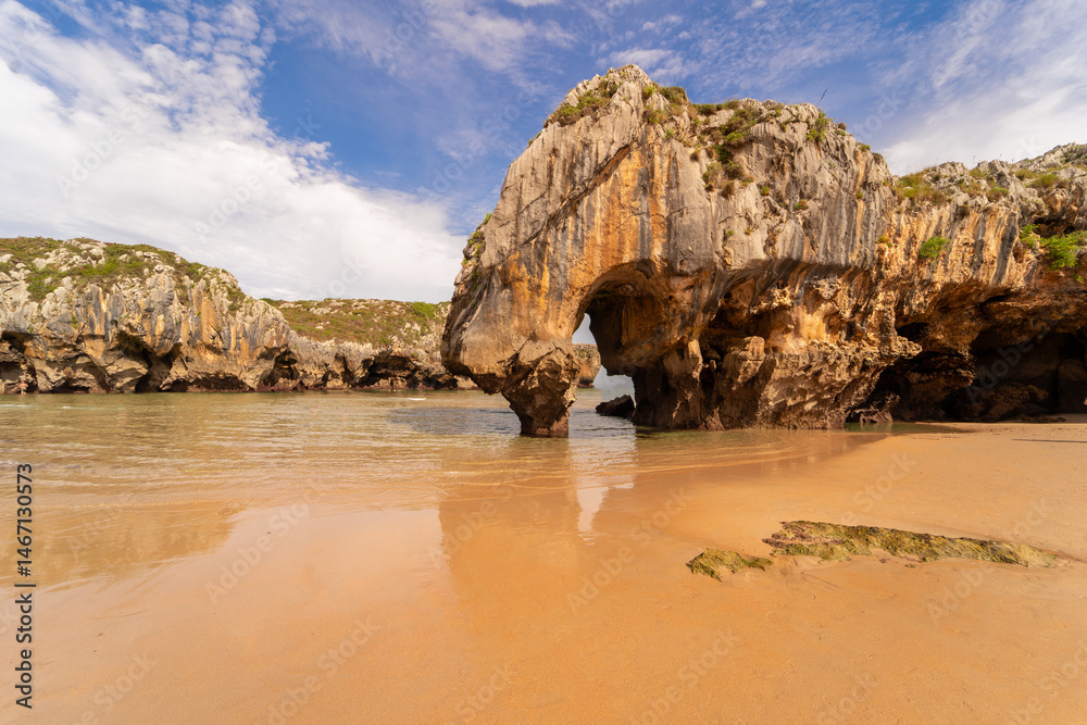 Fototapeta premium Natural rock arch rises from the shallow waters of a golden sandy beach at Playa de Cuevas del Mar in Asturias, Spain.