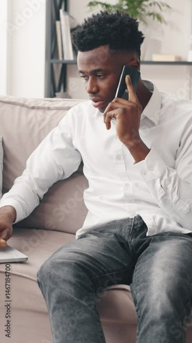 Vertical video with busy african american businessman calling by mobile phone from home, online work with laptop and freelance, part-time job and overworking in weekend, young adult black guy