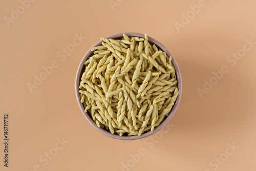 Top view of a bowl of raw italian durum wheat trofie pasta against a beige background .