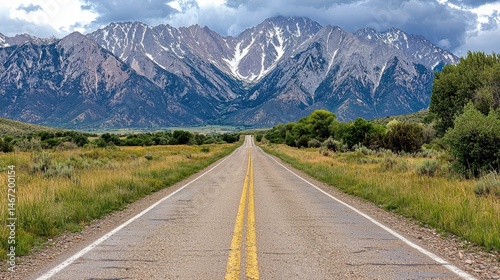 Asphalt road vanishing into the distance towards a majestic snow capped mountain range under a partly cloudy sky. Lush green vegetation flanks the
