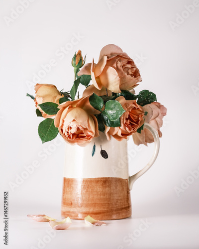 A jug with olive motif full of beautiful orange, pink and yellow roses in bloom against a white background