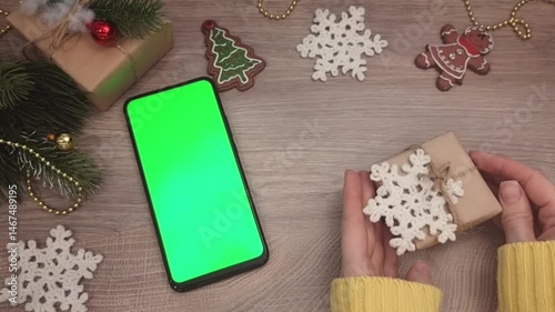 There is a smartphone with a green screen and Christmas decorations on the table , hands appear and put a gift on the table , top view .