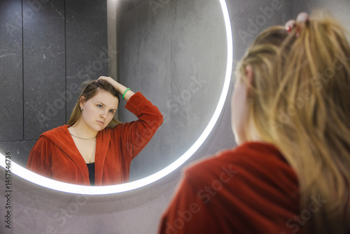 Young woman in a red robe looks at herself in the mirror, lifting her hair with a concerned expression in a modern bathroom with soft lighting.