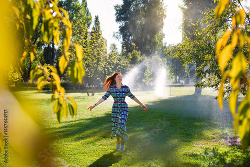 A happy young woman is joyfully walking through a sunny park where sprinklers are actively watering the grass