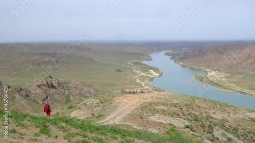 A girl or woman in a checkered shirt with her dog descends to a viewing platform above the Ili River and a nomadic city in Kazakhstan on a sunny spring day. Walks and trips with a pet through natural 