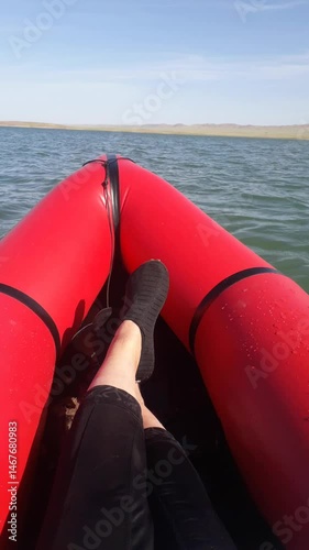 Legs of a girl or woman in aquashoes in a red inflatable packraft swinging on small waves of a lake. Concept about relaxed rest on water, woman in a boat, woman traveler.