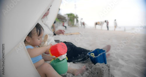 Adorable toddler playing with colorful beach toys under a sunshade on a sandy beach, accompanied by a parent, joyfully scoops sand into a bucket, capturing a moment of summer fun and family bonding.