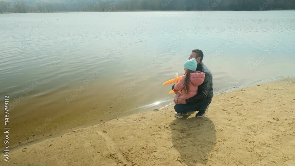 Dad and daughter walking near lake share holding airplane toy on sunny day Dad and daughter walking near lake share holding airplane toy on sunny day