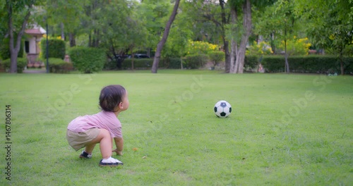 Young Asian toddler sits on green grass in a park, pointing playfully toward a soccer ball. The outdoor scene captures curiosity, play, and early childhood exploration in a peaceful natural
