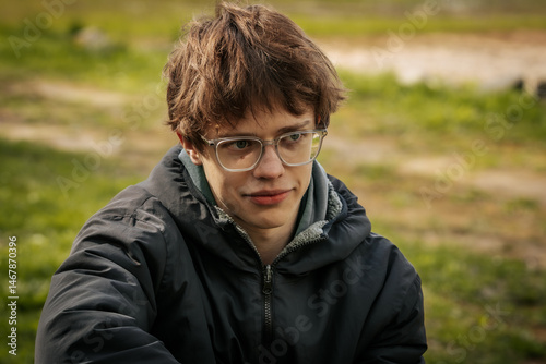 A young man in a black jacket and glasses sits by a wooden table outdoors, appearing thoughtful and relaxed in a grassy natural setting.