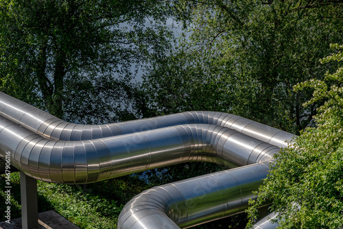 a large, winding pipe for district heating in the sunlight in front of a green tree
