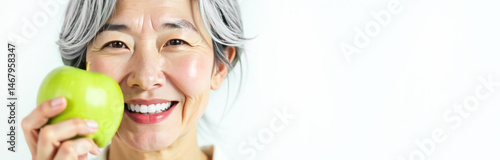 Close-up portrait of a gray-haired adult asian woman holding a green apple and smiling, white beautiful smile on white with space for text.. Dentistry, professional examination, beauty industry