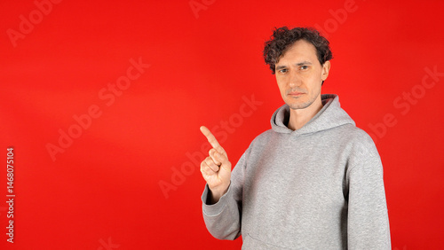 A young man stands against a vibrant red background, pointing with his index finger at an empty space for text, inviting attention and engagement