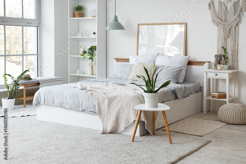 Interior of light bedroom with cozy bed, bedside table, houseplants and shelving unit