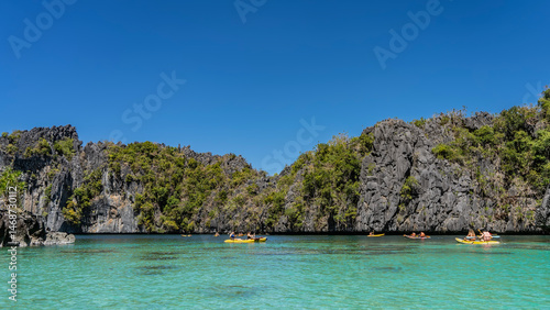 Tourist canoes sail through the beautiful turquoise lagoon surrounded by sheer cliffs. Green tropical vegetation on steep slopes. Clear blue sky. Copy space. Philippines. Palawan. Big Lagoon.