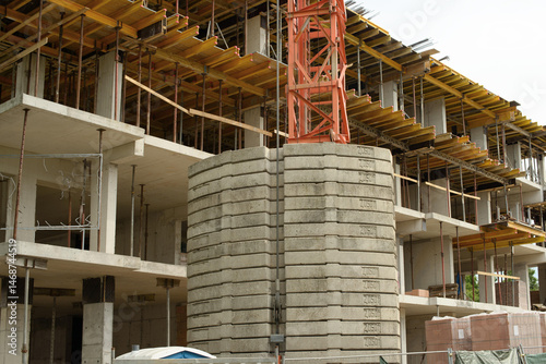 Multi story concrete building under construction featuring yellow formwork scaffolding a red tower crane with concrete counterweights exposed steel reinforcement and open floor slabs on a bright day