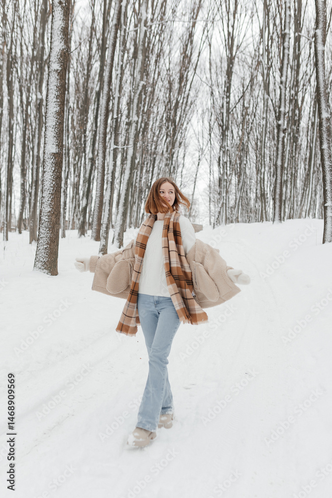 Happy smiling young woman wearing coat, scarf, and mittens enjoying winter weather while walking in snowy winter park. Stylish winter women's clothing.