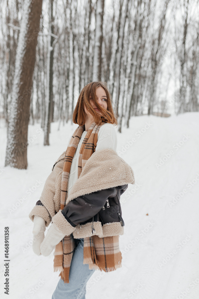 Happy smiling young woman wearing coat, scarf, and mittens enjoying winter weather while walking in snowy winter park. Stylish winter women's clothing.