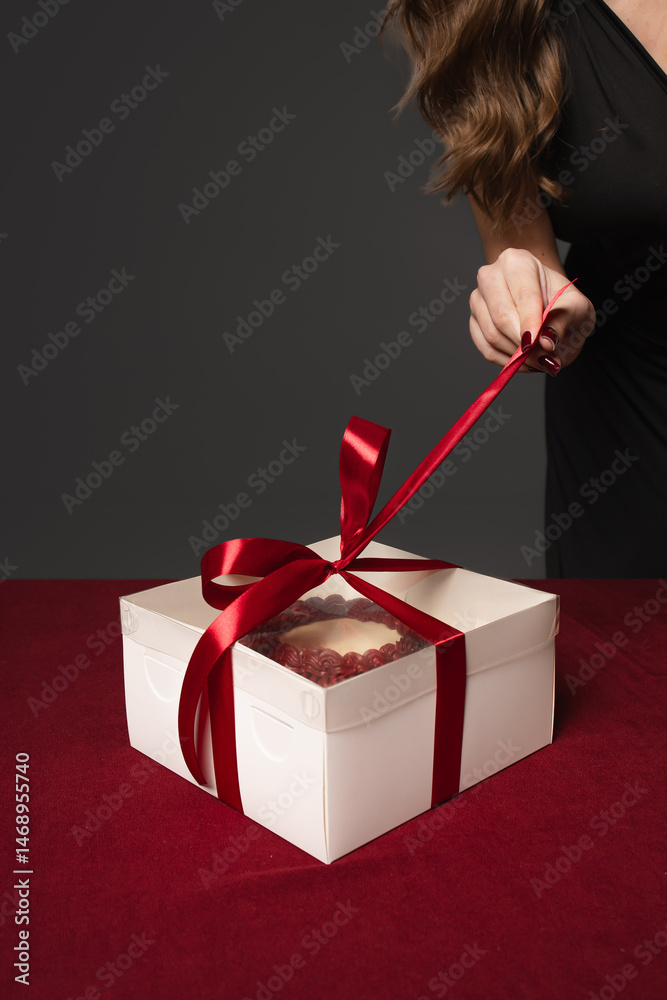 Female hand unties the ribbon on a box with a cake. Close-up of a gift box. The gift is tied with a red ribbon.