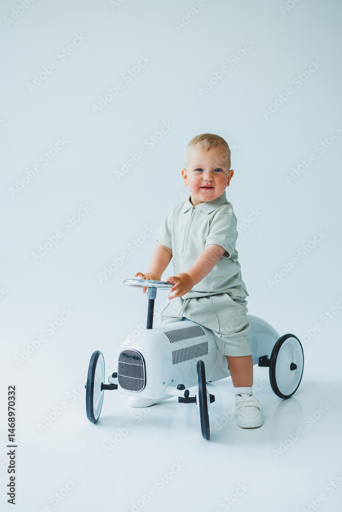 Stylish one-year-old boy in a suit with shorts and a T-shirt rides a car. Toddler boy on a white background with a toy car.
