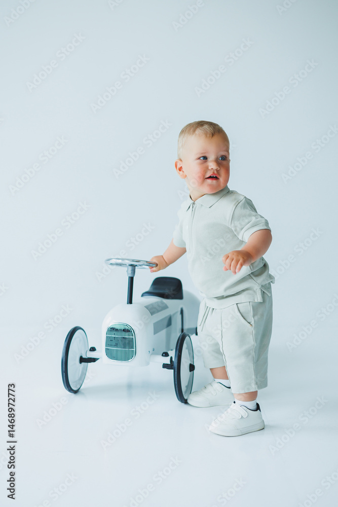 Stylish one-year-old boy in a suit with shorts and a T-shirt rides a car. Toddler boy on a white background with a toy car.