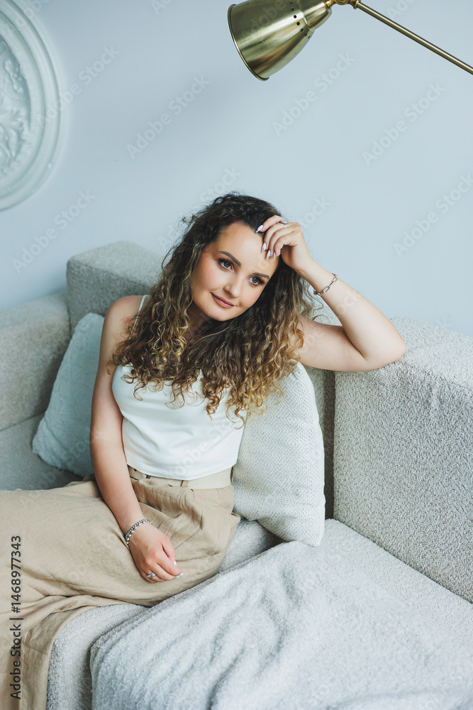 Curly pretty woman sitting in the living room on the sofa and smiling dreamily. Woman in a white top and skirt with a romantic mood sitting in the living room.