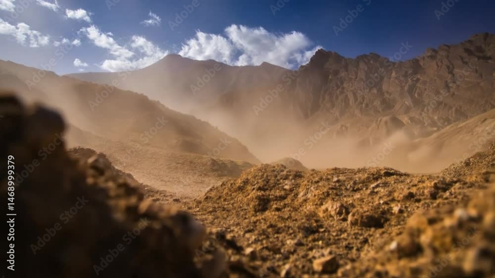 Windy, dusty desert landscape with rocks in the foreground and rugged mountains under a dramatic cloudy sky. Evokes feelings of vastness, desolation, adventure, and the power of nature. Low angle shot