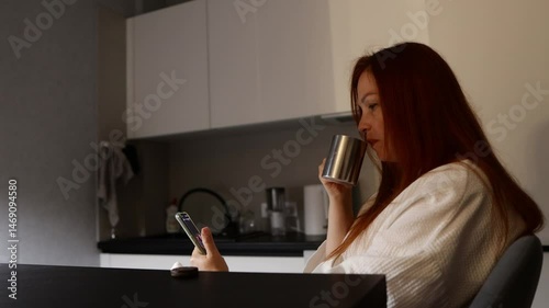 a woman drinks tea in the kitchen late at night and watches social media on a smartphone screen, insomnia and loneliness