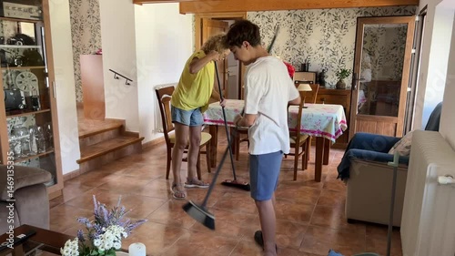 Teenage boy sweeping living room floor as his middle-aged mother assists and supervises during household chores